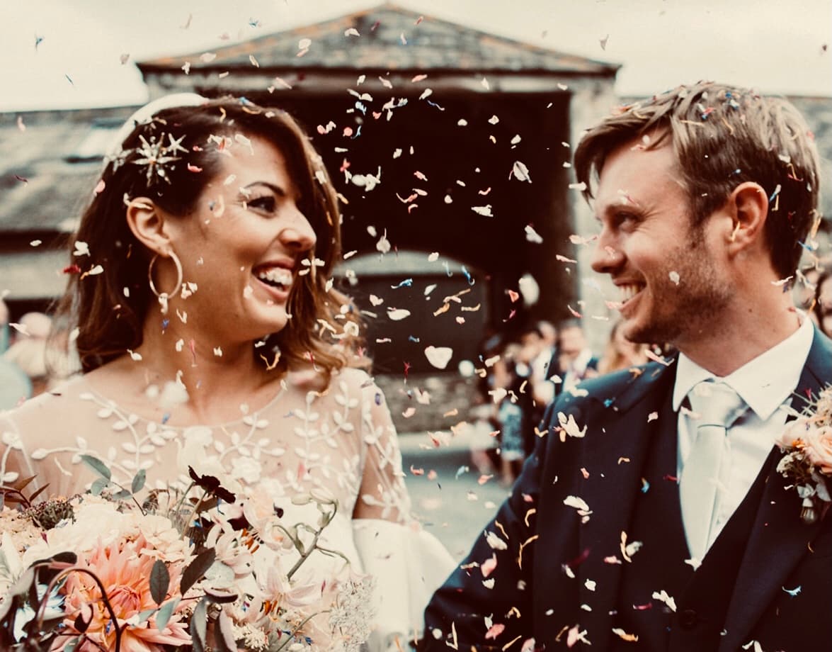 Bride and groom smiling at each other under confetti outside a stone barn