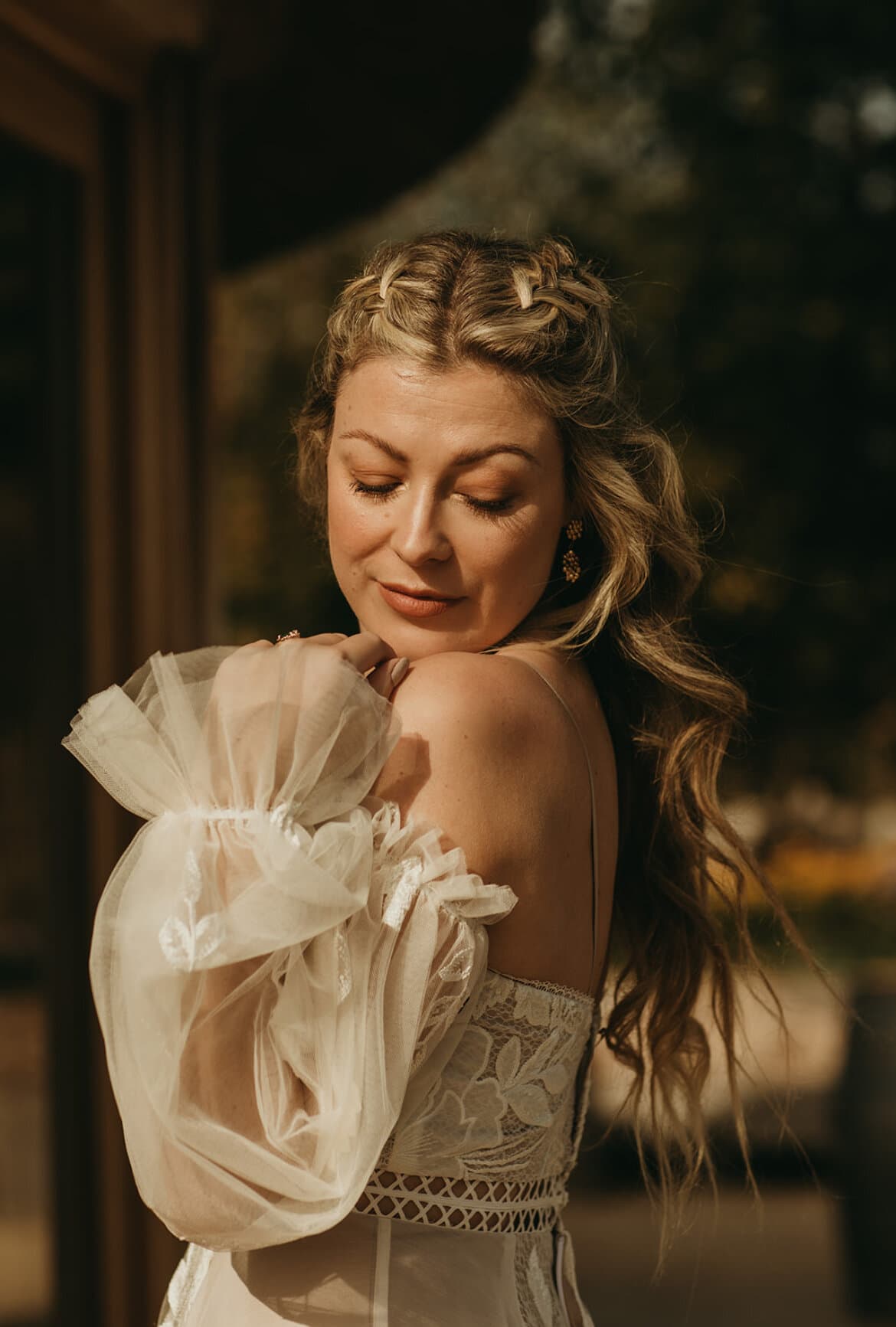 A bride with braided hair and sheer puff sleeves, eyes closed, in warm sunlight