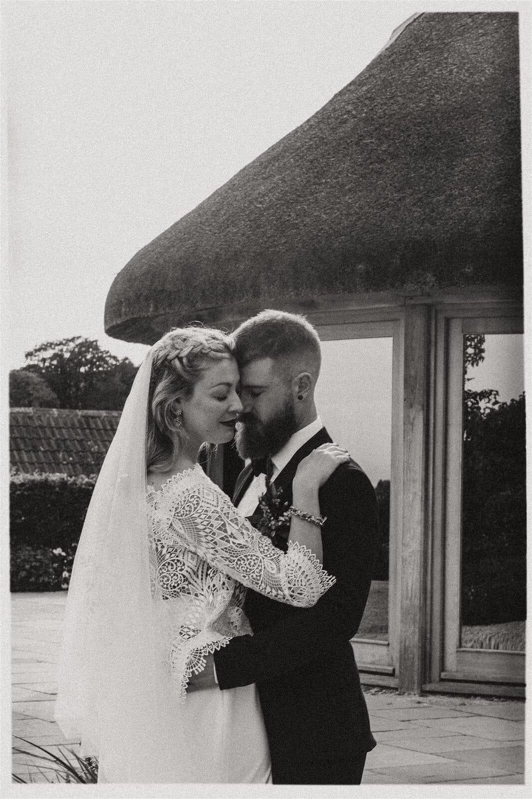 Black and white photo of a couple embracing by a thatched porch